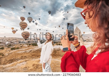 Two friends capture the beauty of Cappadocia, Turkey with their smartphones, surrounded by unique rock formations and hot air balloons. Sharing their adventure on social networks.