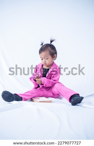 A portrait of a 2 year old girl sitting down, reading colored book on white isolated background. 