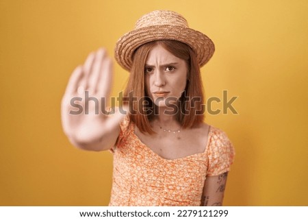 Young redhead woman standing over yellow background wearing summer hat doing stop sing with palm of the hand. warning expression with negative and serious gesture on the face. 
