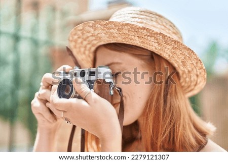 Young redhead woman tourist wearing summer hat using vintage camera at street