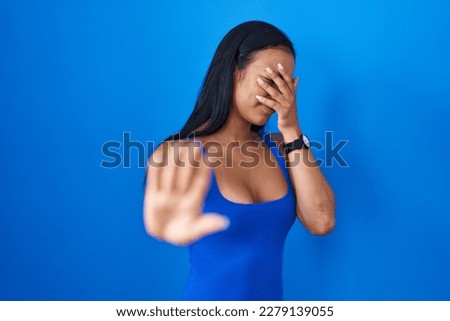 Hispanic woman standing over blue background covering eyes with hands and doing stop gesture with sad and fear expression. embarrassed and negative concept. 
