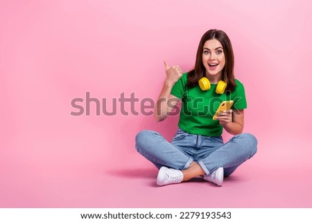 Full length photo of cheerful pretty person sit floor hold telephone direct finger empty space isolated on pink color background