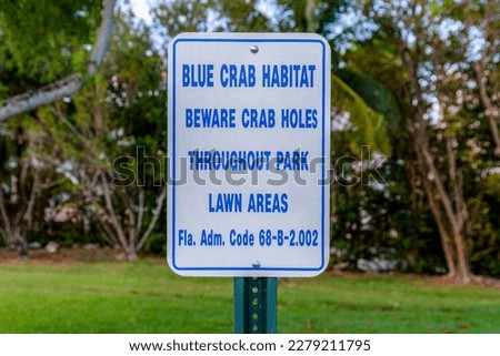Sign post with Blue Crab Habitat Blue Crab Holes Throughout Park Lawn Areas in Miami, Florida. Close-up of a sign post against the lawn and trees in the blurred background.