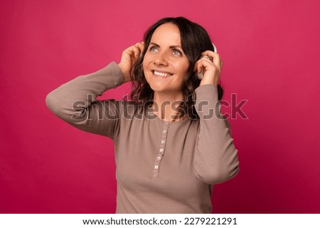 Cheerful mid age woman wearing headphones enjoys listening to the music over pink background.