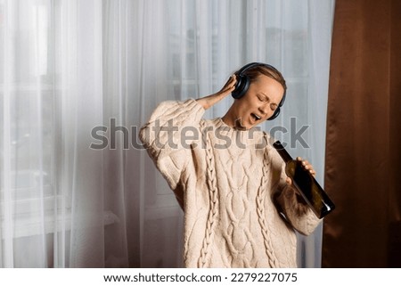 A lovely cheerful woman, a music lover, in a warm loose sweater and headphones listens to music, dances, and sings with a bottle in her hands. Concept of joy and relaxation. Royalty-Free Stock Photo #2279227075