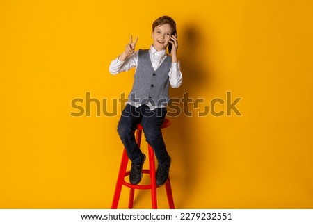 A happy little boy in gray vest and white shirt, neatly dressed, talking on a phone and showing a piece. Isolated on yellow background.