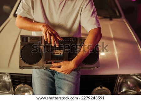 A young man is sitting on a hood of a car playing a cassette on his boombox. Royalty-Free Stock Photo #2279243591
