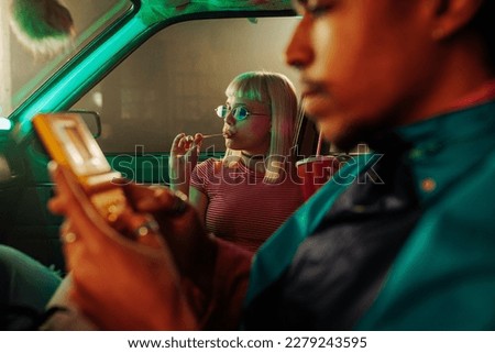 A retro stylish couple is sitting in a car parked. The man is playing a video game, and his girlfriend is next to him with a lollypop.