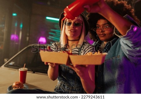 Two young retro fashionable diverse women are outdoors on the hood of their car putting ketchup on their fries.