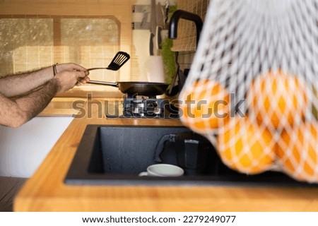 hands cooking with a frying pan inside a camper lined with wood, window light, sink with cups and net with hanging oranges. Designer camper kitchen