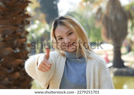 Positive asian girl shows a thumb up looking at the camera in a park. Approval sign or okay.