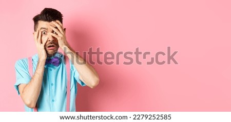Scared and embarrassed nerdy guy in bow-tie covering his eyes, peeking through fingers at something scary, standing on pink background.