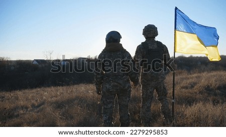Young soldiers in camouflage uniform stands with a waving Ukraine flag at meadow against sunset. Military couple of ukrainian army lifted banner holding hands of each other at field. Dolly shot. Royalty-Free Stock Photo #2279295853