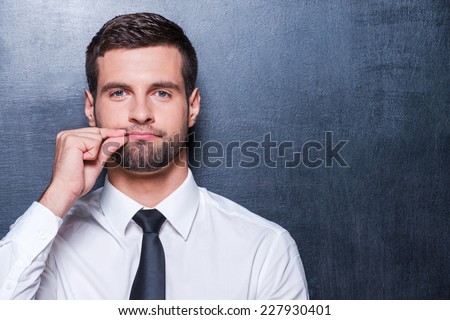 Keep silence! Handsome young man in formalwear showing a sign of closing mouth and looking at camera while standing against blackboard Royalty-Free Stock Photo #227930401