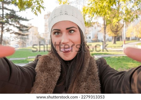Young beautiful long haired brunette with white woolen hat taking a selfie photo in the public park