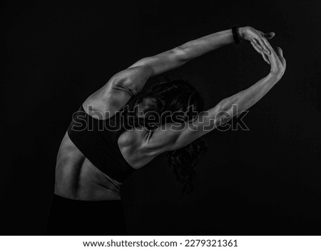Athletic fitness strong woman in sport bra and black leggings doing side bend stretches exercise on dark shadow background with empty space. Back side view portrait
