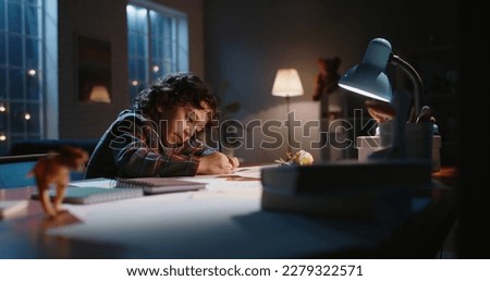 Funny little asian kid drawing at home. Boy with curly hair drawing with pencils on paper in the evening, learning art, having fun at home - hobby concept 
