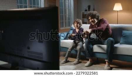 cheerful father and son playing video game at home. Father And Son Sitting On Sofa In Lounge Playing Video Game. Happy family concept.