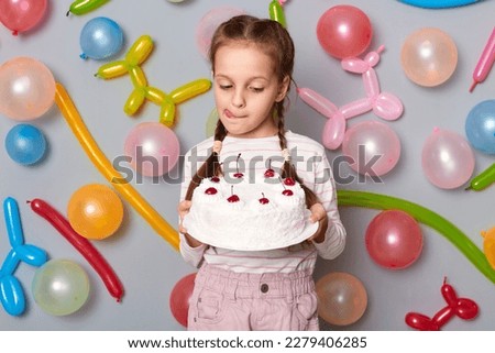 Portrait of hungry little girl with braids wearing casual clothing standing against gray wall with colorful balloons, looking at beautiful tasty cake, showing tongue out.