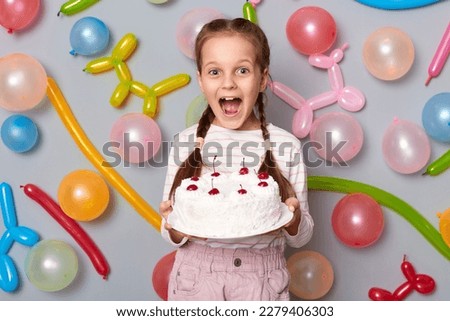 Image of amazed extremely happy little girl with braids wearing casual clothing posing isolated over gray background with balloons, holding birthday cake.