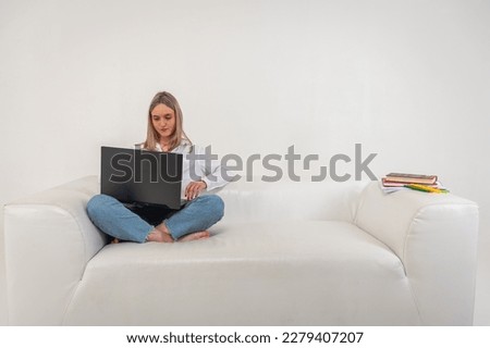 young beautiful girl is sitting on a laptop on the sofa, isolated on a light background, the girl is shopping or chatting online in social networks, working remotely. A place for writing