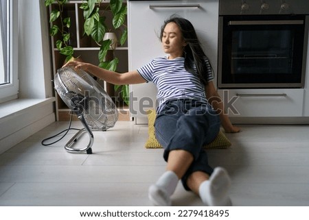 Overheating in homes. Tired overheated young Asian woman sitting on floor in kitchen near electric fan, cooling down at home during extreme summer heat, staying cool without air conditioning Royalty-Free Stock Photo #2279418495