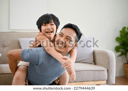 Joyful and happy Asian dad playing with his little son in the living room, piggyback, spending fun time together at home. Royalty-Free Stock Photo #2279444639