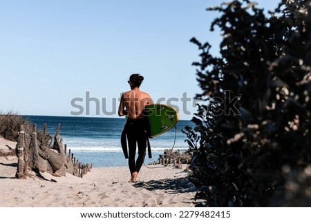 Surfer with wetsuit getting ready for a surf session. Boy walking on the beach with a green surfboard under the arm. Back point of view. Ocean waves and surf. Surfing in Australia.