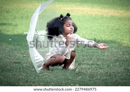Happy smiling African girl with black curly hair with umbrella under raindrops fall while playing outdoor green park, beautiful kid playing outside garden on rainy day, cute child playing in the rain Royalty-Free Stock Photo #2279486975