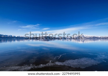 The lake on a clear winter day with fresh blue sky, mirror lake surface, and snowy mountains in the distance. Lake Kussharo in Hokkaido, Japan. Royalty-Free Stock Photo #2279495995