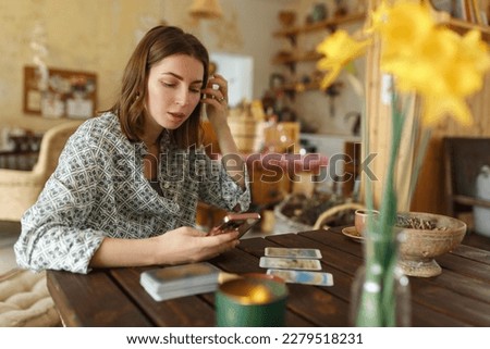 Beautiful young woman is guessing on cards with tarot, runes on wooden table and uses an online app in phone to interpret divination, home interior,candles and dry herbs.. Royalty-Free Stock Photo #2279518231