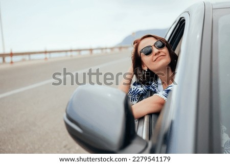 Happy smiling young woman passenger showing thumb up hand, drinking takeaway coffee and looking out car window. Enjoying car trip, summer vacation