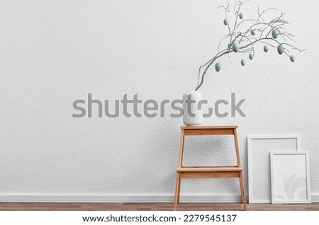 Vase with tree branch, Easter eggs and blank picture frames near light wall