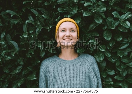 Photo of friendly smiling attractive woman on background of green hedge. Happy confident woman in front of green leaves wall. Thoughtful person outdoors at autumn season.