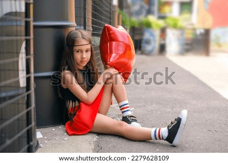 A pre-adolescent girl is sitting in a city courtyard holding a balloon in her hand and looking into the camera with a languid look.  A girl in the image of a hippie Royalty-Free Stock Photo #2279628109