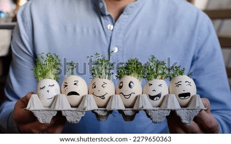 Funny easter eggs with different painted mines in a cartoon holding by hands of man. Green watercress sow and grow in eggs in spring. Banner.