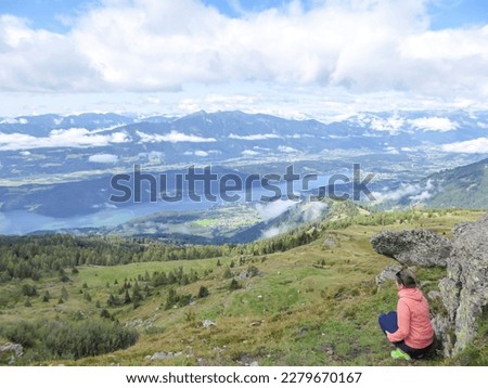 A woman in orange hoodie squatting under a rock and enjoying the view on the Millstaetter lake from Granattor in Austrian Alps. The distant lake is surrounded by high mountains. Few clouds above. Royalty-Free Stock Photo #2279670167