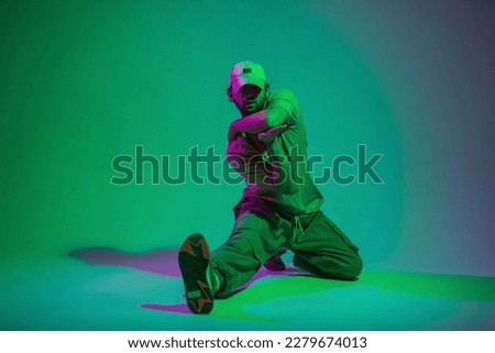 Fashionable handsome professional dancer man with a cap in fashion clothes sits and dances in a creative color studio with cyan and pink lights