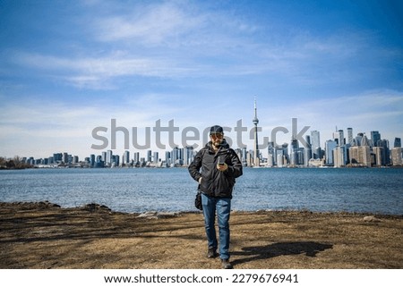 man holds a cellphone in hand on the coast of toronto island park with toronto skyline on background