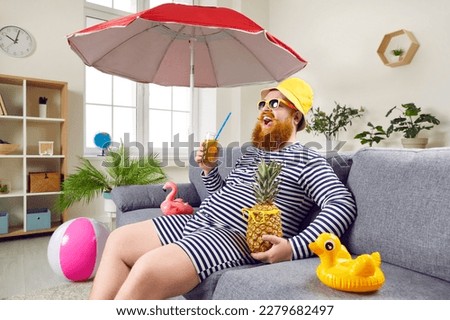 Happy bearded man in classic bathing suit sitting on sofa at home. Excited eccentric man in sunglasses and panama hat sitting under umbrella drinking cocktail enjoying summer vacation in living room Royalty-Free Stock Photo #2279682497