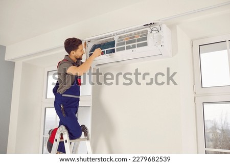 Professional electrician man with screwdriver maintaining, cleaning modern air conditioner indoors. Young technician standing on a ladder in the room repairing or installing air conditioner. Royalty-Free Stock Photo #2279682539