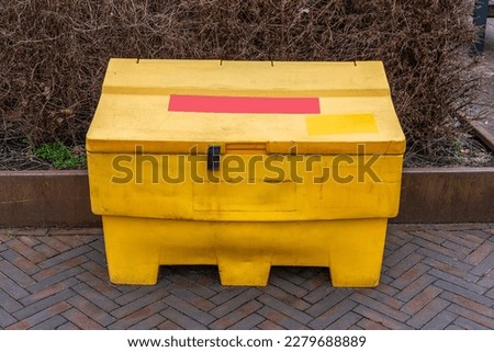 yellow grit salt box standing on the street in front of a thicket