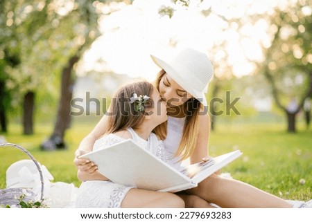 Mom and little daughter are looking at a family album with photos in the summer in the garden. Mom kisses her daughter. Family values