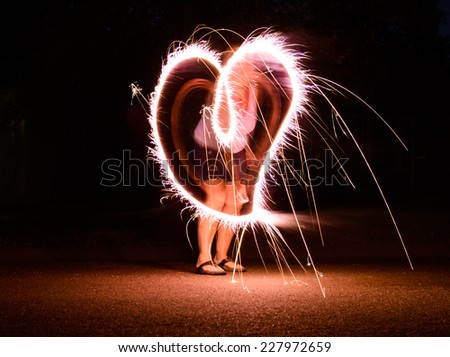A woman celebrates Independence Day with a sparkler, tracing the outline of a heart against a black background. Shot is taken with slow shutter speed. 