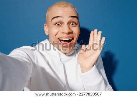Close up young dyed blond man of African American ethnicity wear white hoody doing selfie shot pov on mobile cell phone spread hand isolated on plain dark royal navy blue background studio portrait