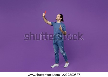 Full body young man in basic blue t-shirt doing selfie shot on mobile cell phone post photo on social network show v-sign isolated on plain purple background studio portrait. People lifestyle concept