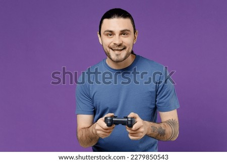 Young smiling cheerful amazed fun cool man 20s wearing basic blue t-shirt hold in hand play pc game with joystick console isolated on plain purple background studio portrait. People lifestyle concept