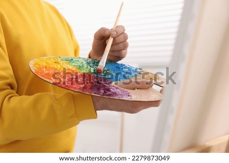 Man mixing paints on palette with brush near canvas in studio, closeup