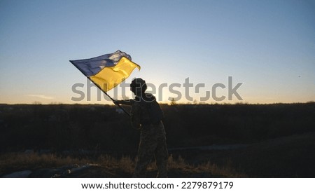 Military man in uniform waving flag of Ukraine against background of sunset. Male ukrainian army soldier lifting national banner at hill in honor of the victory against russian aggression. Rear view.