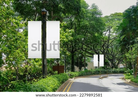 Blank vertical advertising banners on street lampposts; double hanging posters by the road, against lush green trees and plants. For OOH out of home template mock up. Royalty-Free Stock Photo #2279892581